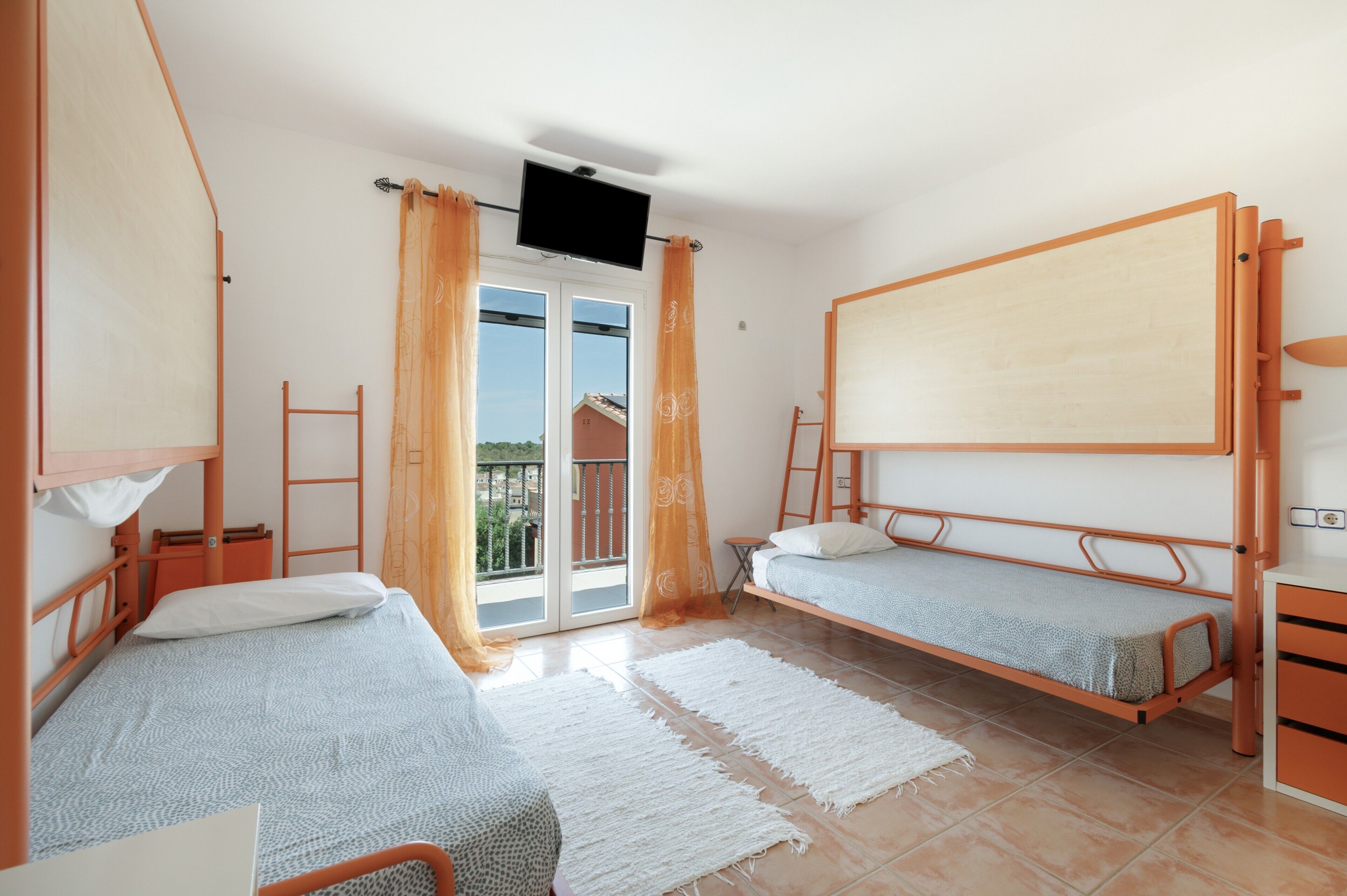Un dormitorio con dos literas, un televisor montado en la pared, un balcón con cortinas anaranjadas y una puerta corredera de vidrio.