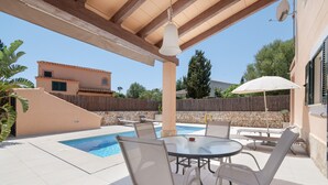 An outdoor pool area with a table and chairs, a hanging light fixture, and a stone wall in the background. - Villas 2meet La Yuca – Holiday Home with Private Pool in Cala Romántica, Mallorca (Romàntica)