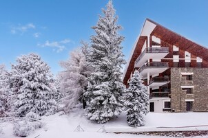 A modern building with a sloped roof and multiple balconies, surrounded by snow-covered trees.