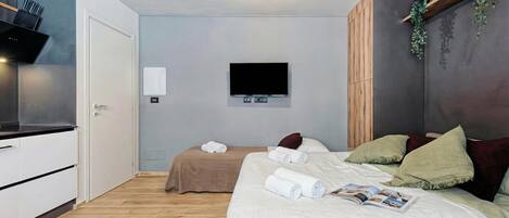 A modern hotel room with a wooden floor, a bed with white bedding, a flat-screen TV mounted on the wall, and a small kitchen area with a microwave and a coffee machine.