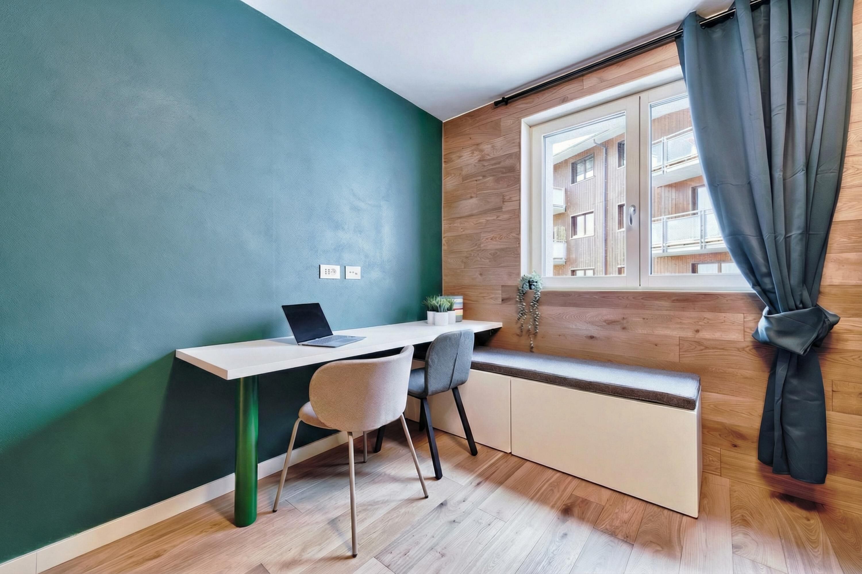 Ein kleines Zimmer mit Schreibtisch, Stuhl, Fenster mit Vorhängen und Blick auf Gebäude.