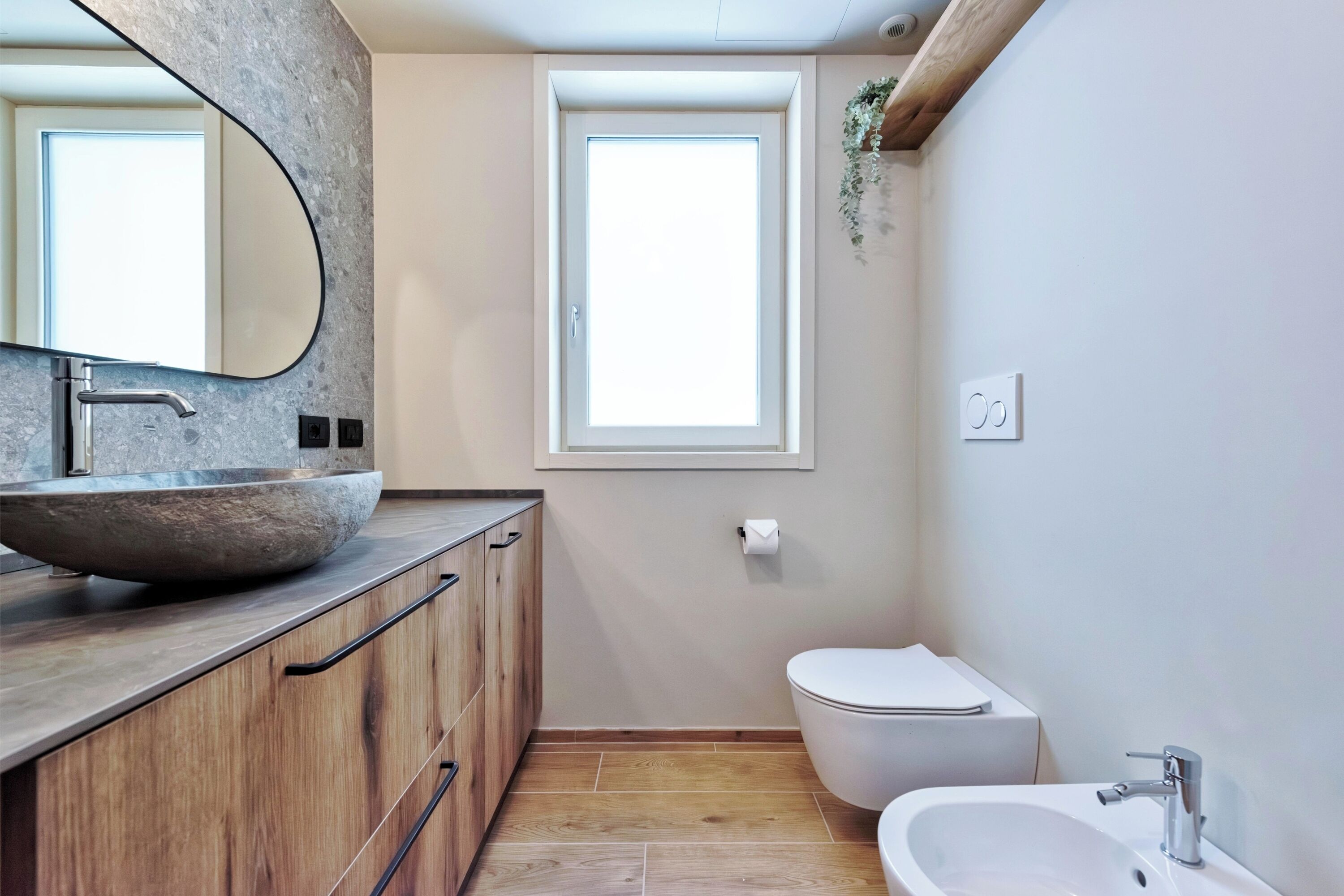 Un bagno moderno con un lavabo ovale, mobili in legno, un water e una finestra che lascia entrare luce naturale.