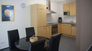 Una cocina compacta con armarios de madera, un microondas y una estufa. Hay una mesa de comedor con sillas y un reloj en la pared.