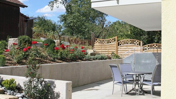 Un balcón con mobiliario exterior, un jardín con flores rojas y un cielo azul despejado.