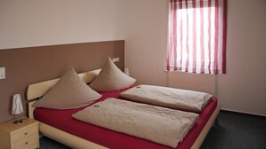 Una cama doble con colcha roja y dos almohadas, una mesita de noche con lámpara y una ventana con cortinas a rayas.
