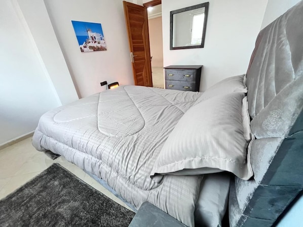 Un lit bien fait, avec une couette et des oreillers gris, une tête de lit bleu foncé, une petite commode en bois et un tableau encadré au mur.