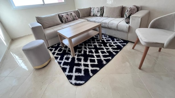 Ein modernes Wohnzimmer mit einem Ecksofa, einem Couchtisch, einem grauen Pouf und einem gemusterten Teppich.