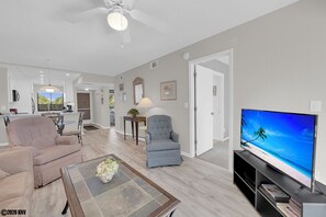 A modern living room with a flat-screen TV, a beige sofa, a blue armchair, and a coffee table with a vase. - 2B/2B Fairview Isles 104, Walk to Beach, Heated Pool! (Fort Myers Beach)