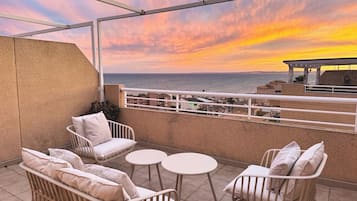 Une terrasse dotée de meubles en osier blanc et offrant une vue sur la mer au coucher du soleil.