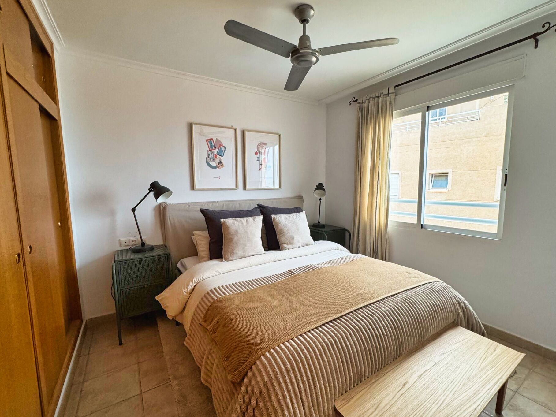 Une chambre à coucher comprenant un lit, un ventilateur de plafond, deux tables de chevet avec des lampes, une fenêtre avec des rideaux et des œuvres d’art accrochées au mur.