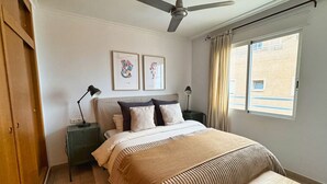 Ein Schlafzimmer mit einem Bett, Deckenventilator, zwei Nachttischen mit Lampen, einem Fenster mit Vorhängen und Kunstwerken an der Wand.