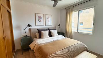 Une chambre à coucher comprenant un lit, un ventilateur de plafond, deux tables de chevet avec des lampes, une fenêtre avec des rideaux et des œuvres d’art accrochées au mur.