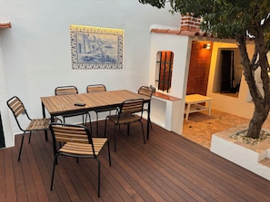 Um deck de madeira com mesa e cadeiras, uma parede revestida de azulejos com um detalhe decorativo e uma pequena árvore.