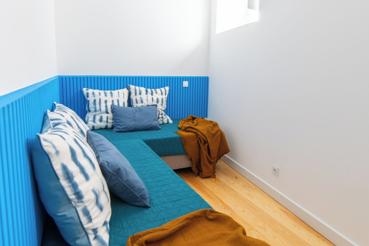 Una habitación pequeña y moderna con una pared azul de acento, una cama individual con almohadas estampadas, una lámpara colgante azul y un ventanal que proporciona luz natural.