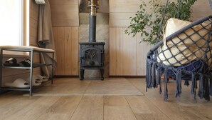 Una habitación acogedora con piso de madera, un estufa de leña negra, una silla con cojín de macramé y un estante de metal con zapatos.