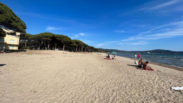 En sandstrand med solbadere og gående mennesker, en klar blå himmel og en bygning med træer i nærheden.