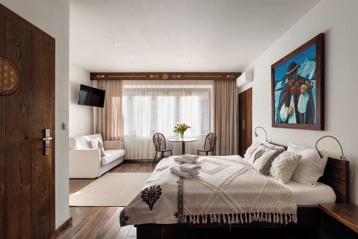 Habitación de hotel con cama, sofá, televisión, un cuadro y ventana con cortinas.