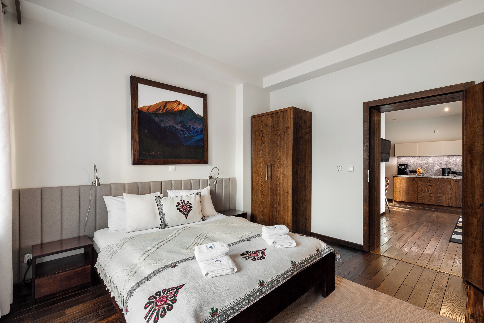 A bedroom with a bed, a nightstand, a wooden wardrobe, a painting, and a view into the kitchen.