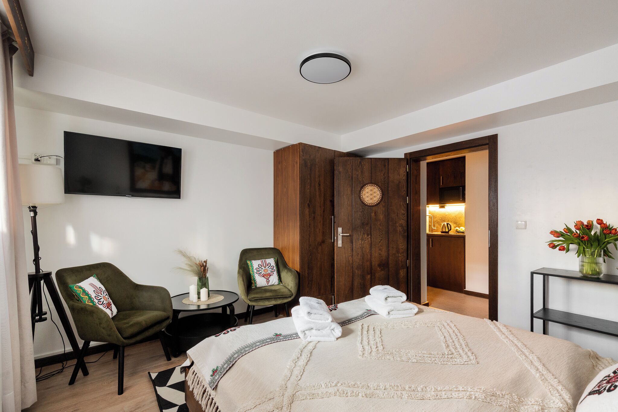 A hotel room with a bed, two armchairs, a small table, a TV, a wardrobe, and a bathroom visible through an open door.
