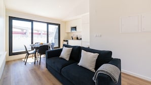 A modern living room with a dark blue sofa, two white cushions, and a grey throw blanket. There is a dining area with a table and chairs, and a kitchen area with a microwave and cabinets. - Alcântara 2 (Lisbon)