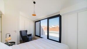 A modern bedroom with a large bed, a chair, a desk, and a view of the city through a large window. - Alcântara 2 (Lisbon)