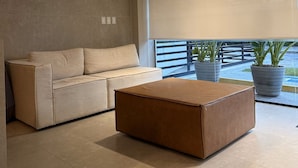A modern living room with a beige sofa, a brown ottoman, and large windows with blinds. - Flat Cabo Branco Aurea 50m do mar 602 (João Pessoa)