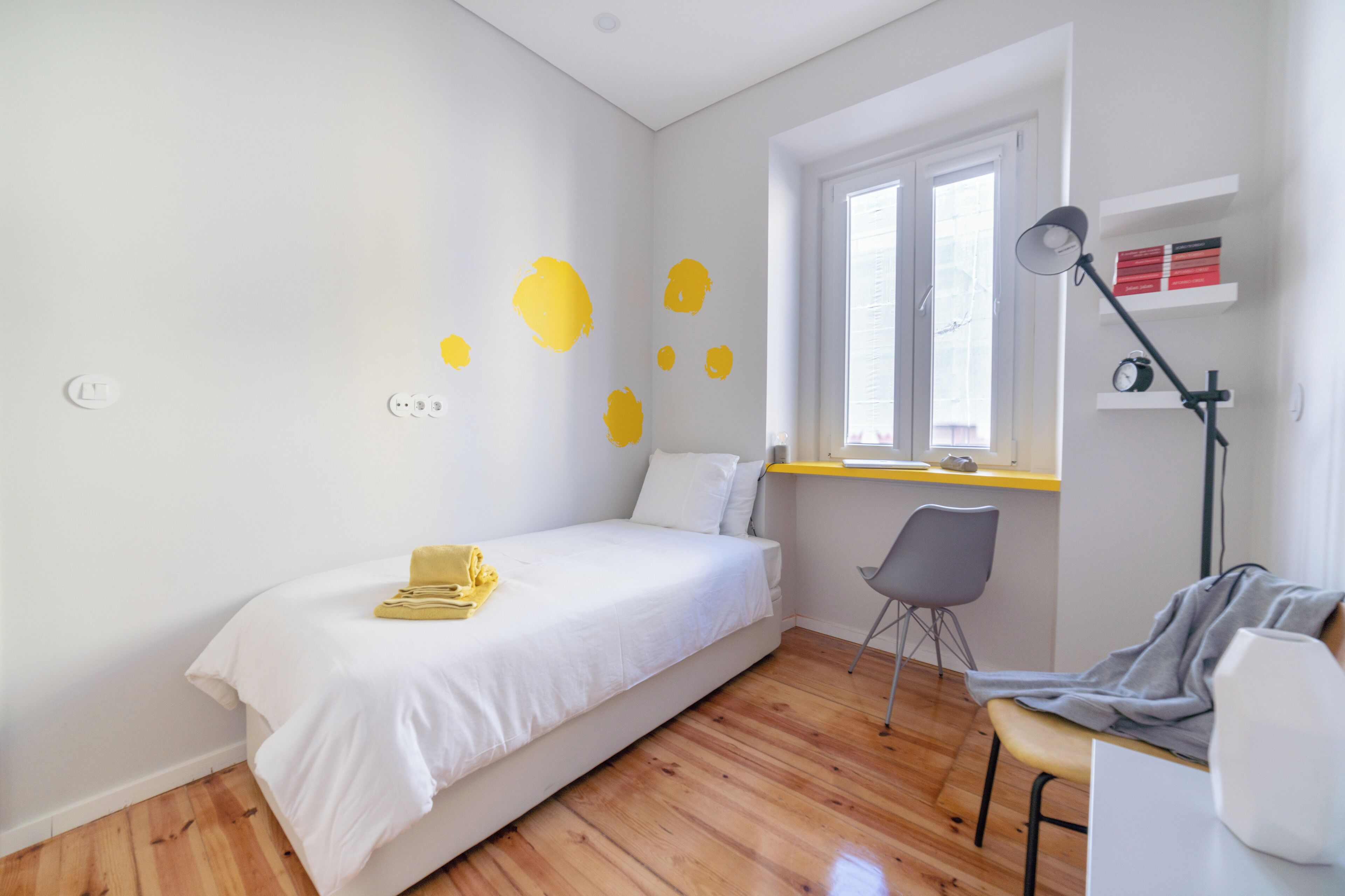A bedroom with a bed, a desk, a chair, a window, and a shelf with books and a clock.
