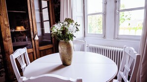 Une table ronde blanche, sur laquelle est posé un vase de fleurs, entourée de chaises blanches, dans une pièce dotée de portes en bois et d’une fenêtre donnant sur un paysage verdoyant.