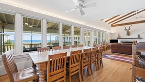 Un comedor amplio con una larga mesa de madera y sillas, ventanas grandes que ofrecen vistas a la playa y un ventilador de techo.