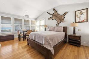 Een slaapkamer met een groot bed, een houten vloer, een bureau met stoelen en een raam met jaloezieën.