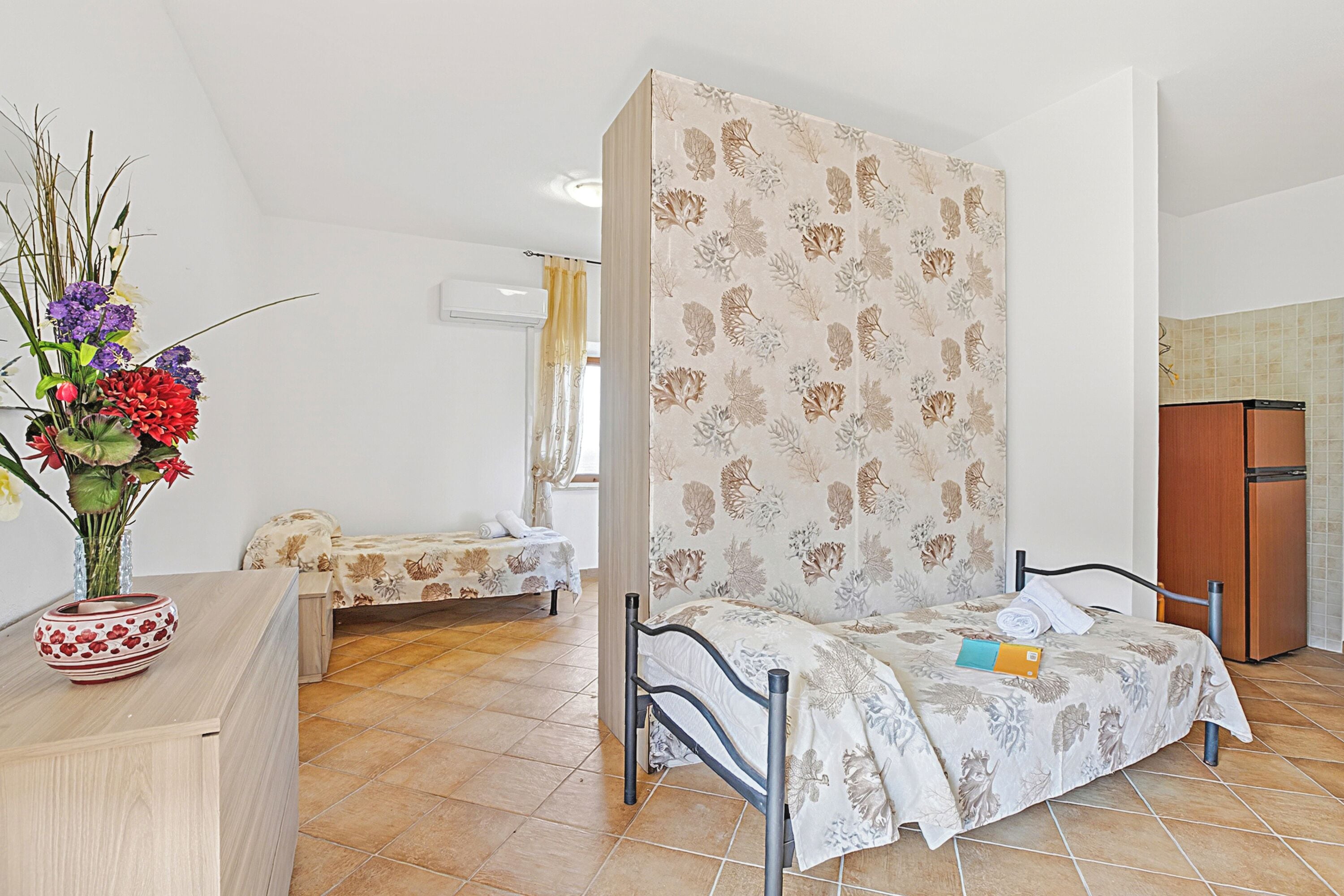 Une chambre à coucher avec un lit, une table de chevet ornée d’un vase, une petite table et une fenêtre avec des rideaux.