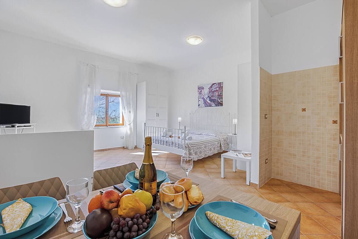 A room with a bed, a dining table with plates of fruit and a bottle, a TV, and a window with curtains.