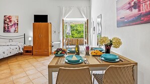 A bedroom with a bed, a wooden wardrobe, a small table with a bottle, and a dining area with a table set for two. - Residence Argentiera Apartments (Sassari)