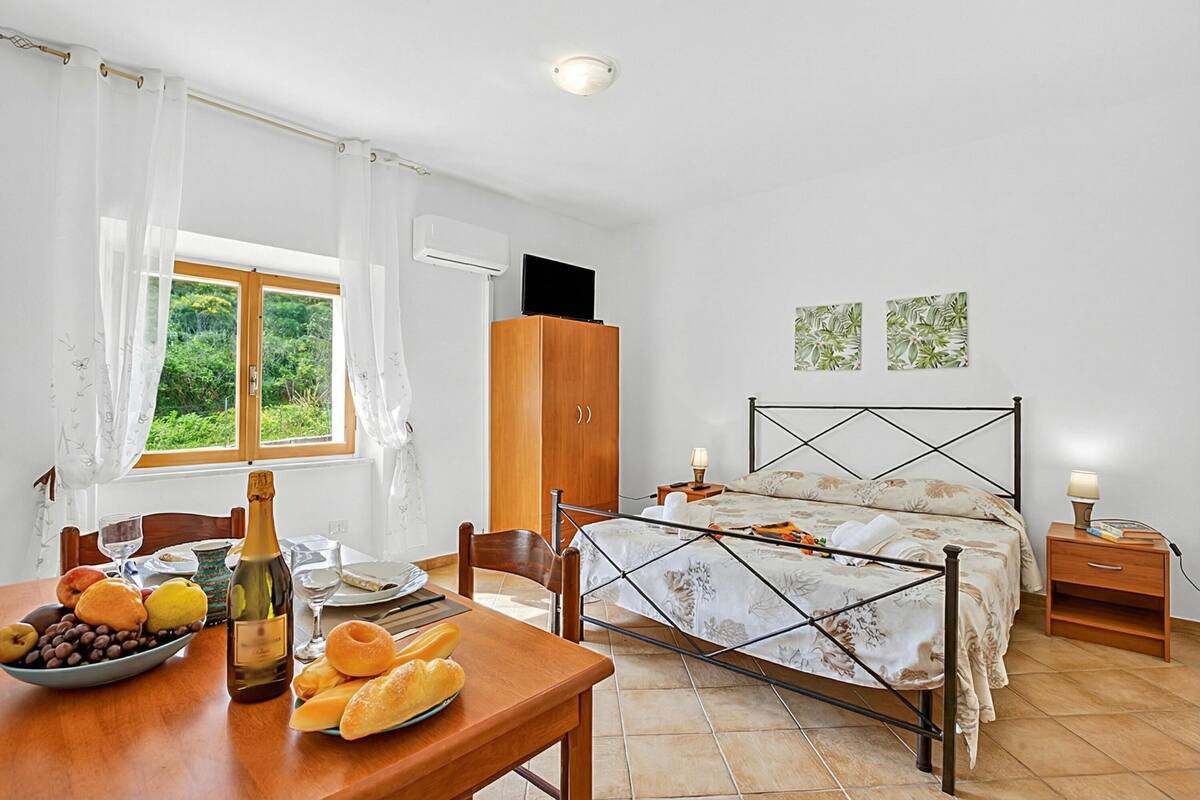 A bedroom with a bed, a wooden wardrobe, a bedside table with a lamp, a window with curtains, and a table with fruits and a bottle.