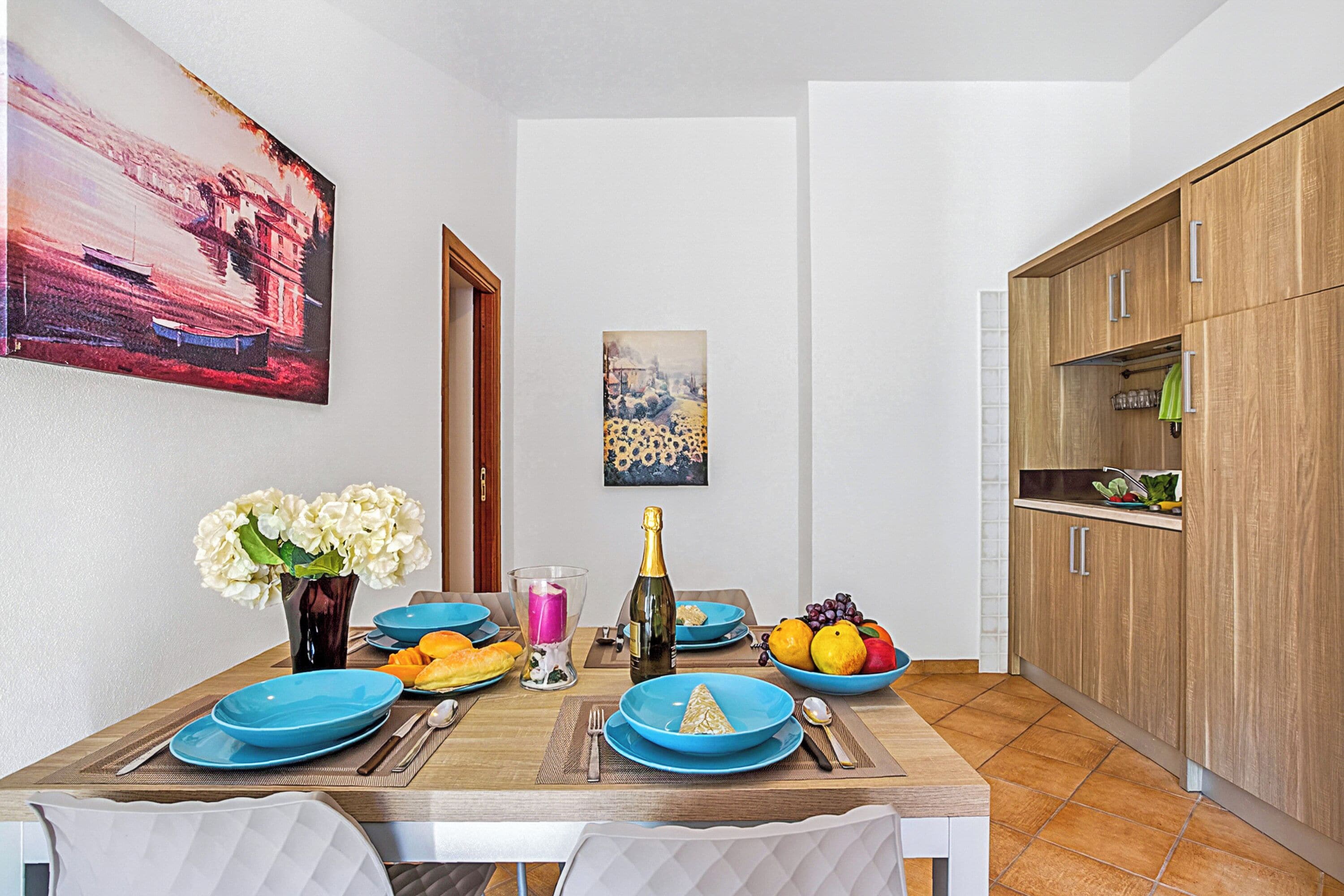 Een eethoek met een houten tafel voor twee personen, een bloevas, een fles champagne en een schaal fruit.