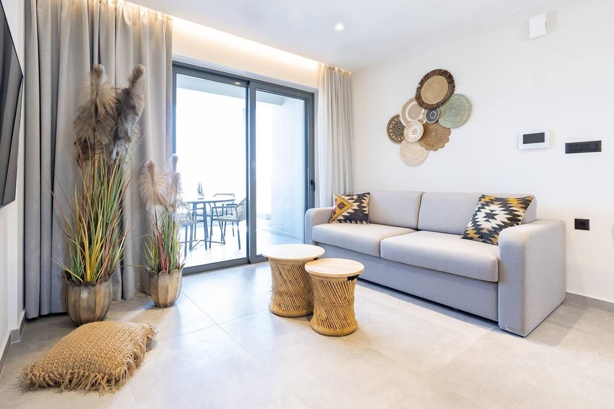 Uma sala moderna com um sofá cinzento, duas mesas laterais redondas de fibra natural, uma grande planta em vaso e vista para uma área externa através de uma porta de vidro.