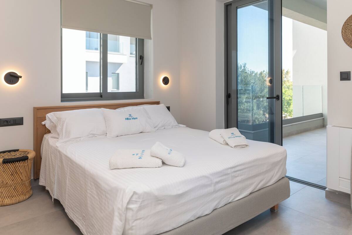 Quarto de hotel moderno com uma cama grande, duas mesinhas de cabeceira, janela com persianas e porta para varanda.