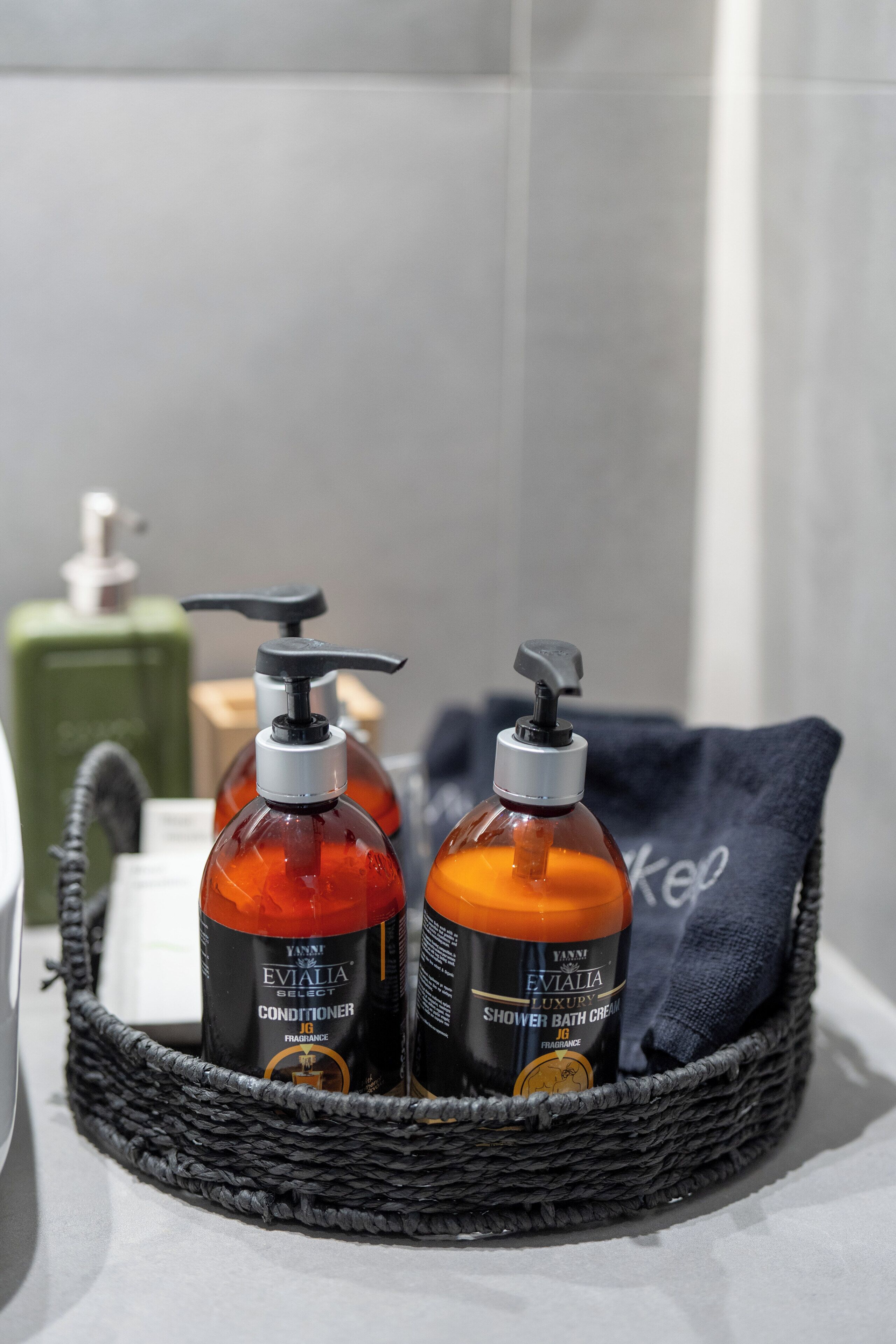 A bathroom with a wicker basket containing two bottles of Yanni Evialia Select conditioner and shower bath cream, a green soap dispenser, and a blue towel.
