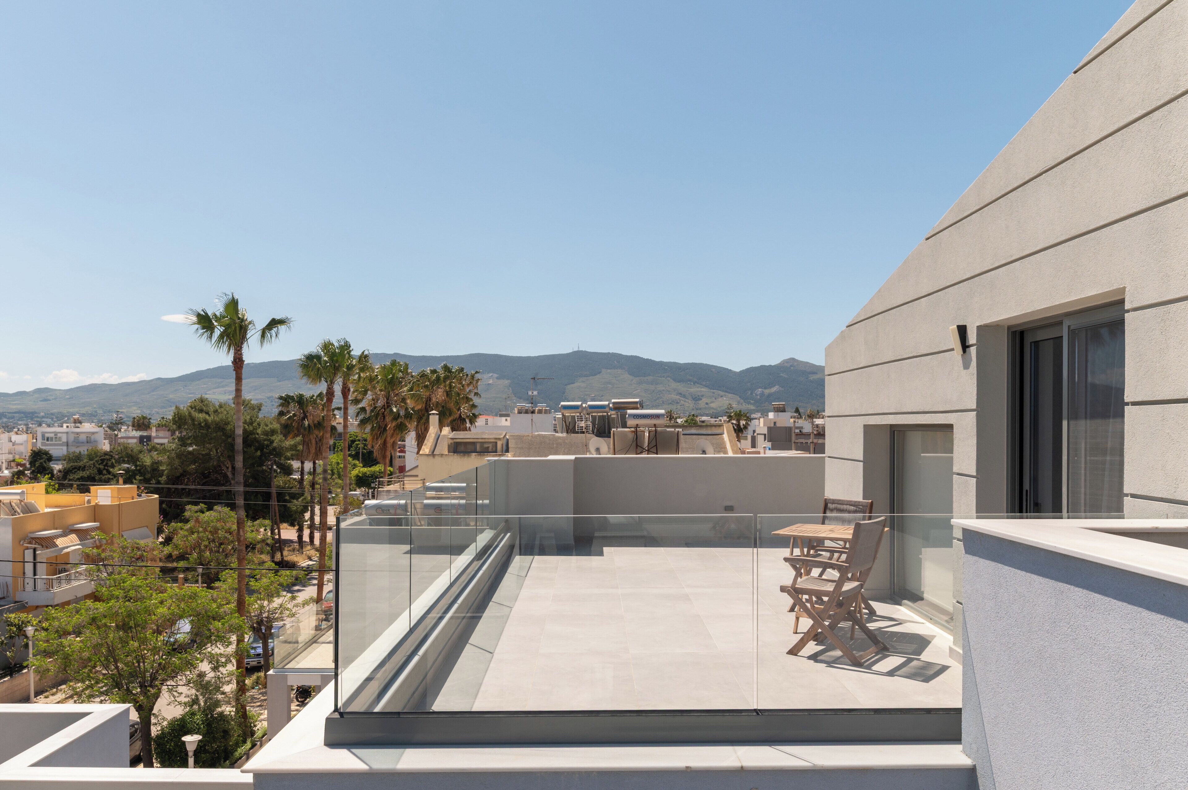 A rooftop terrace with a glass balustrade, a table and chairs, and a view of the cityscape and mountains.
