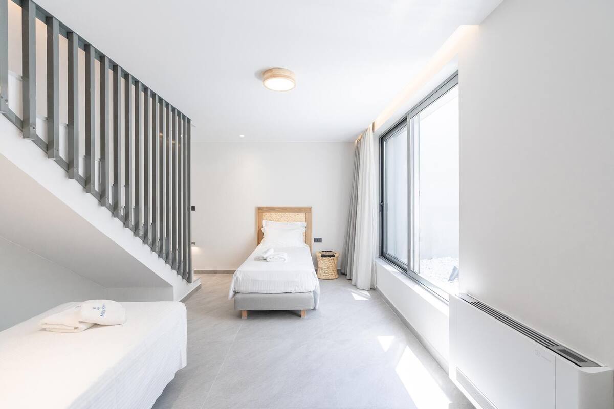 Quarto de hotel moderno com cama, um pequeno espaço para sentar, janela com cortinas e escada com corrimão.