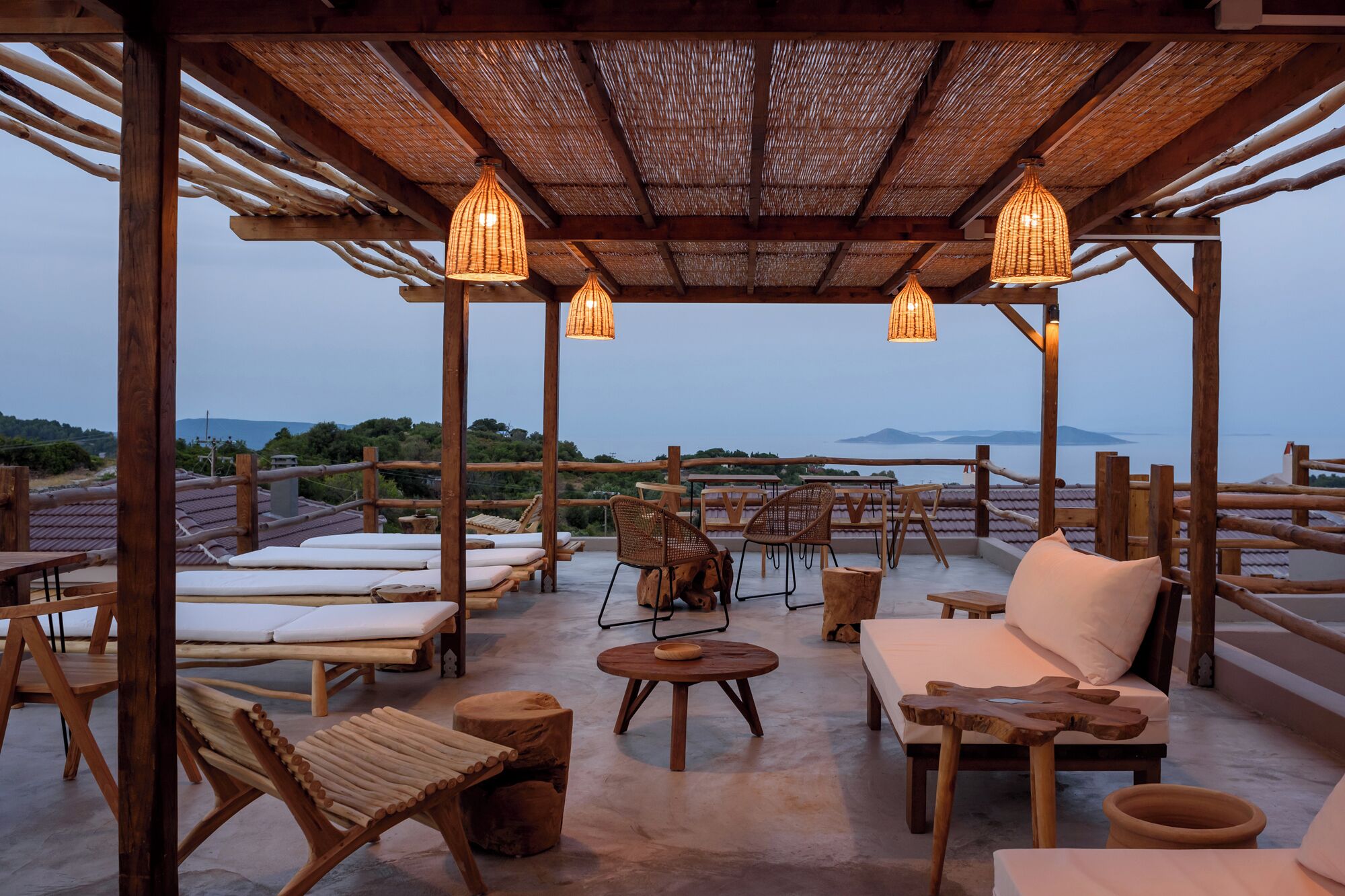 A covered outdoor seating area with wooden furniture, woven light fixtures, and a view of the sea.