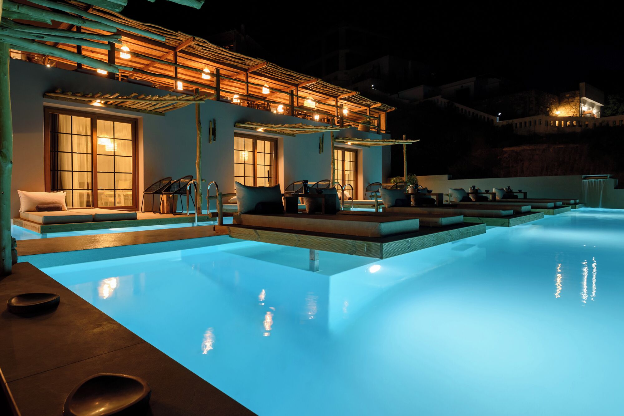 Vue nocturne d’un espace piscine avec une terrasse en bois, des chaises longues et un coin salon couvert offrant une vue sur la piscine.