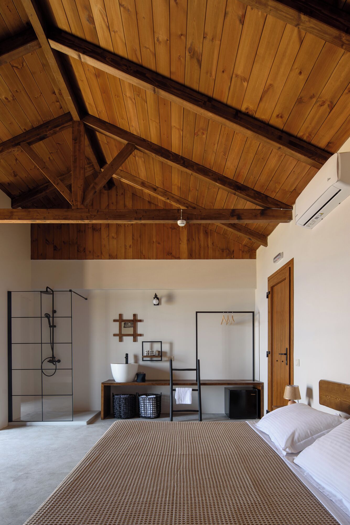 A bedroom with a wooden ceiling, a large bed, a bedside table, a mirror, a wardrobe, and an air conditioning unit.