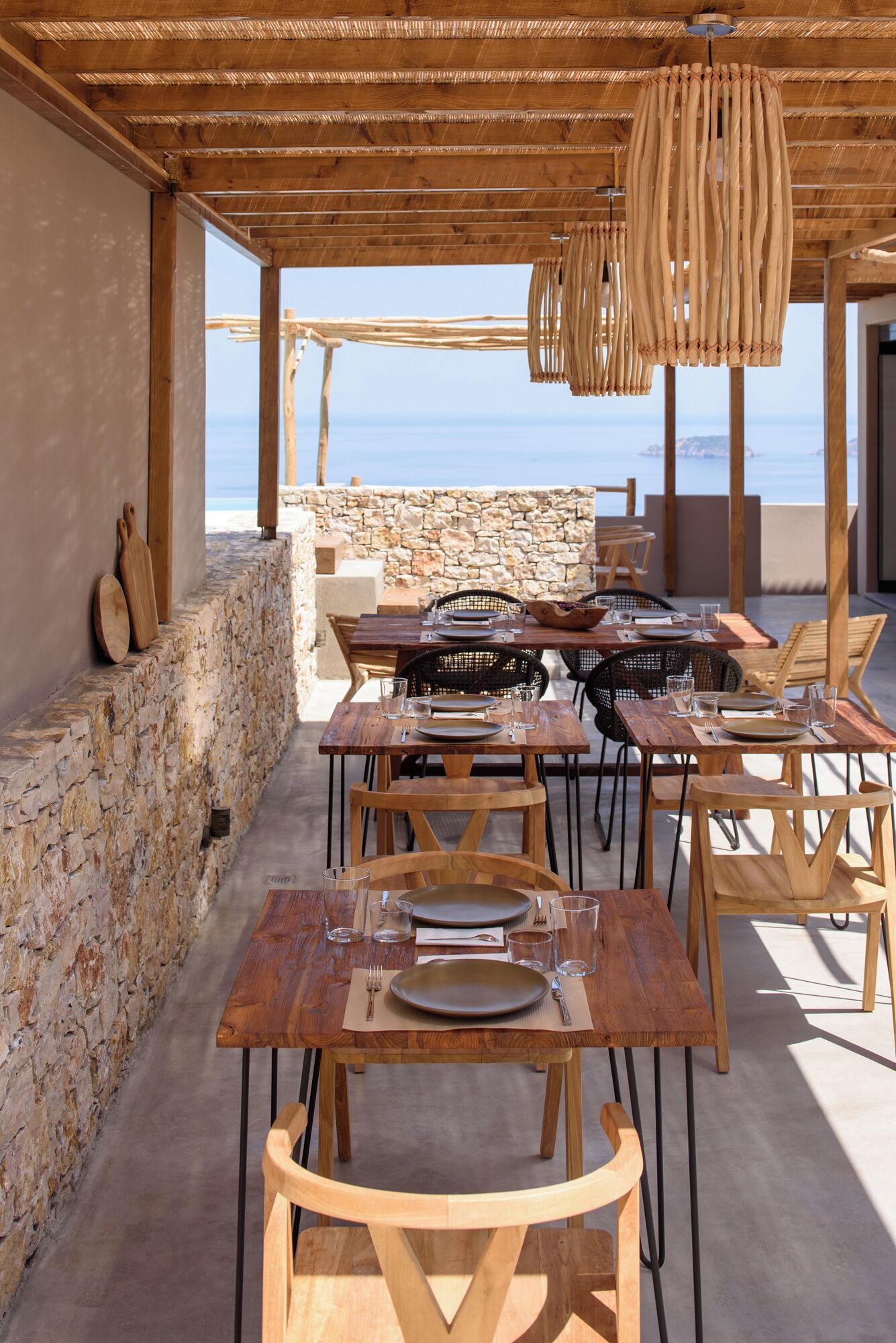 An outdoor dining area with wooden tables and chairs, stone walls, and a view of the sea.