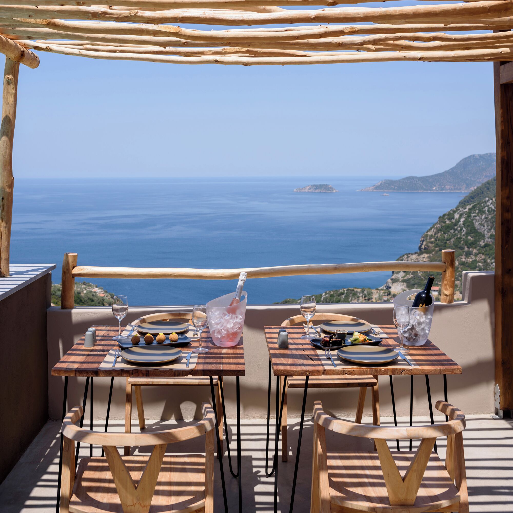 A dining setup with two tables, each with plates, cutlery, and a bottle of wine, overlooking a coastal view.