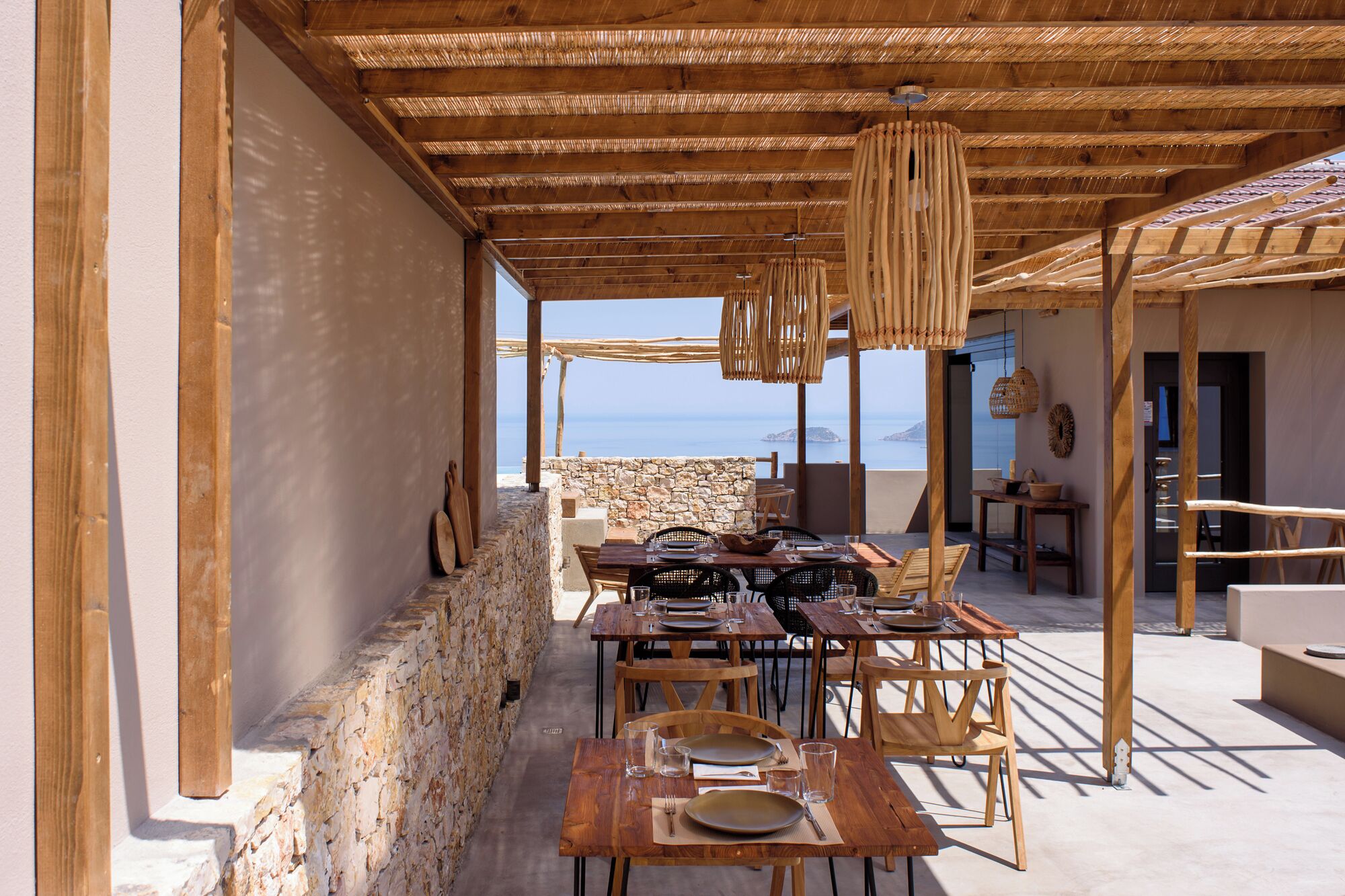 Un espacio para comer al aire libre con mesas y sillas de madera, paredes de piedra y vistas al mar.