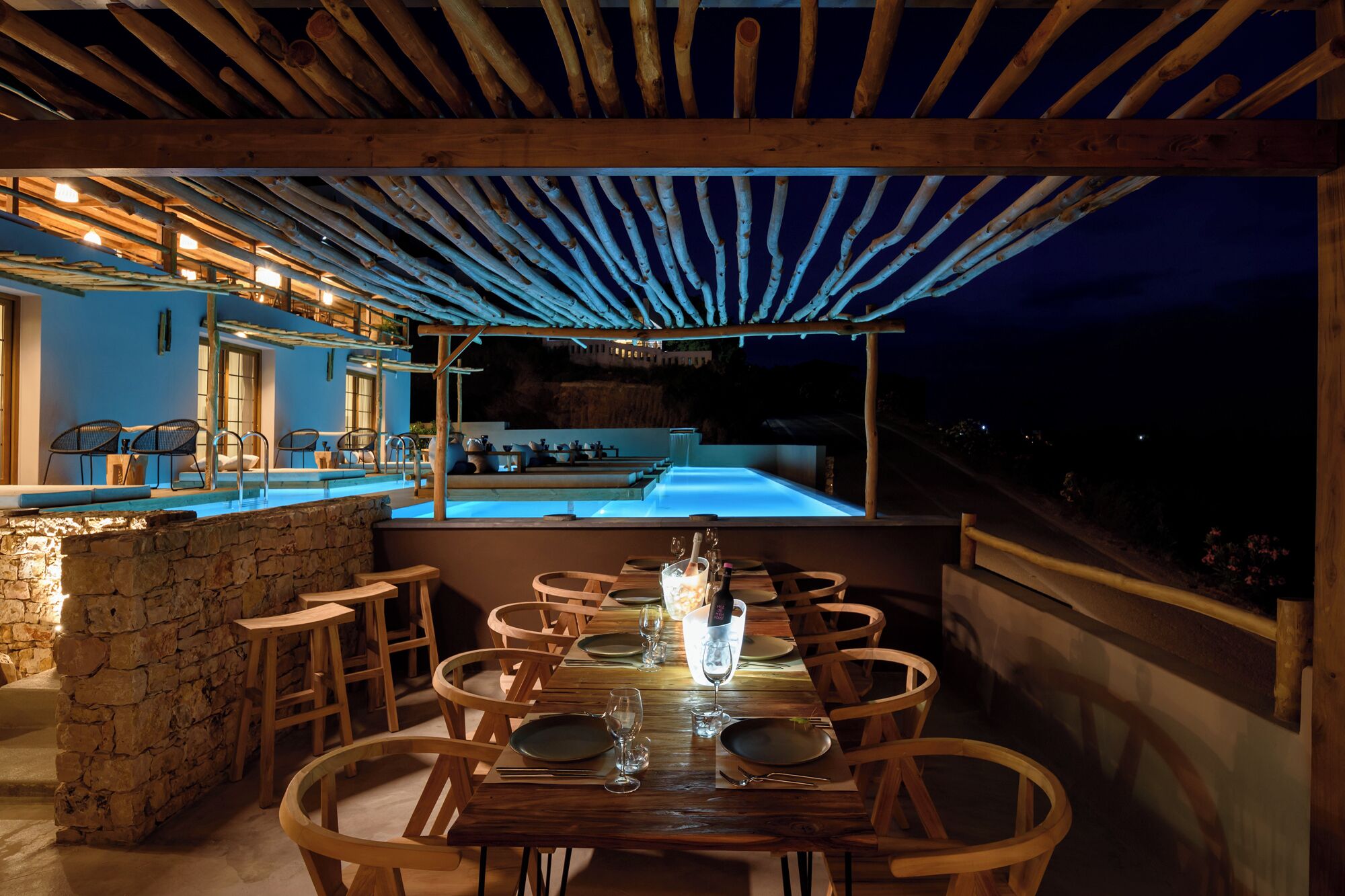 An outdoor dining area with a pool and a bar, featuring wooden furniture and a stone wall.