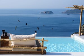 Un área de descanso junto a la piscina con vistas al mar y a las islas, muebles de madera y un cojín blanco.