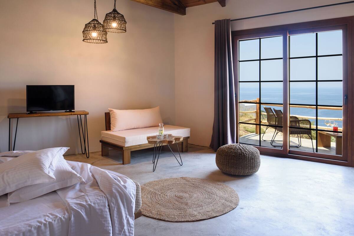 A bedroom with a bed, a sofa, a TV, a table with a glass, and a view of the sea through large windows.