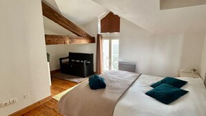 Un dormitorio con techo inclinado, vigas de madera, cama con ropa blanca y almohadas turquesa, mesita de noche y ventana con cortinas.